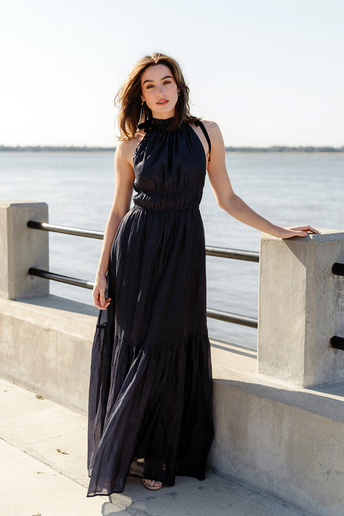 A woman standing on a pier wearing a navy maxi dress with a high neckline, sleeveless design, and a flowy skirt with a playful ruffle hem.