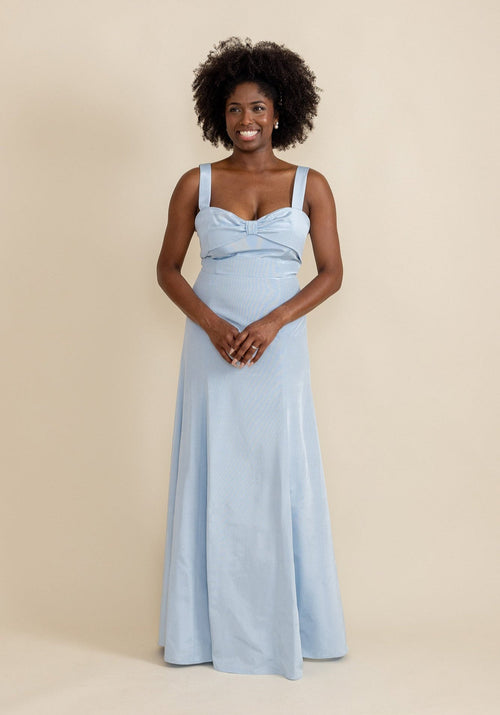 A woman standing, wearing a blue dress with a sweetheart neckline and bow detail on a flare skirt.